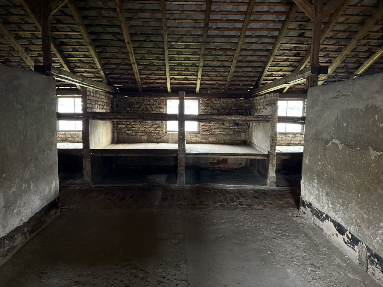 Pohled do prázdné ubikace s dřevěnými pryčnami a okny v Auschwitz-Birkenau