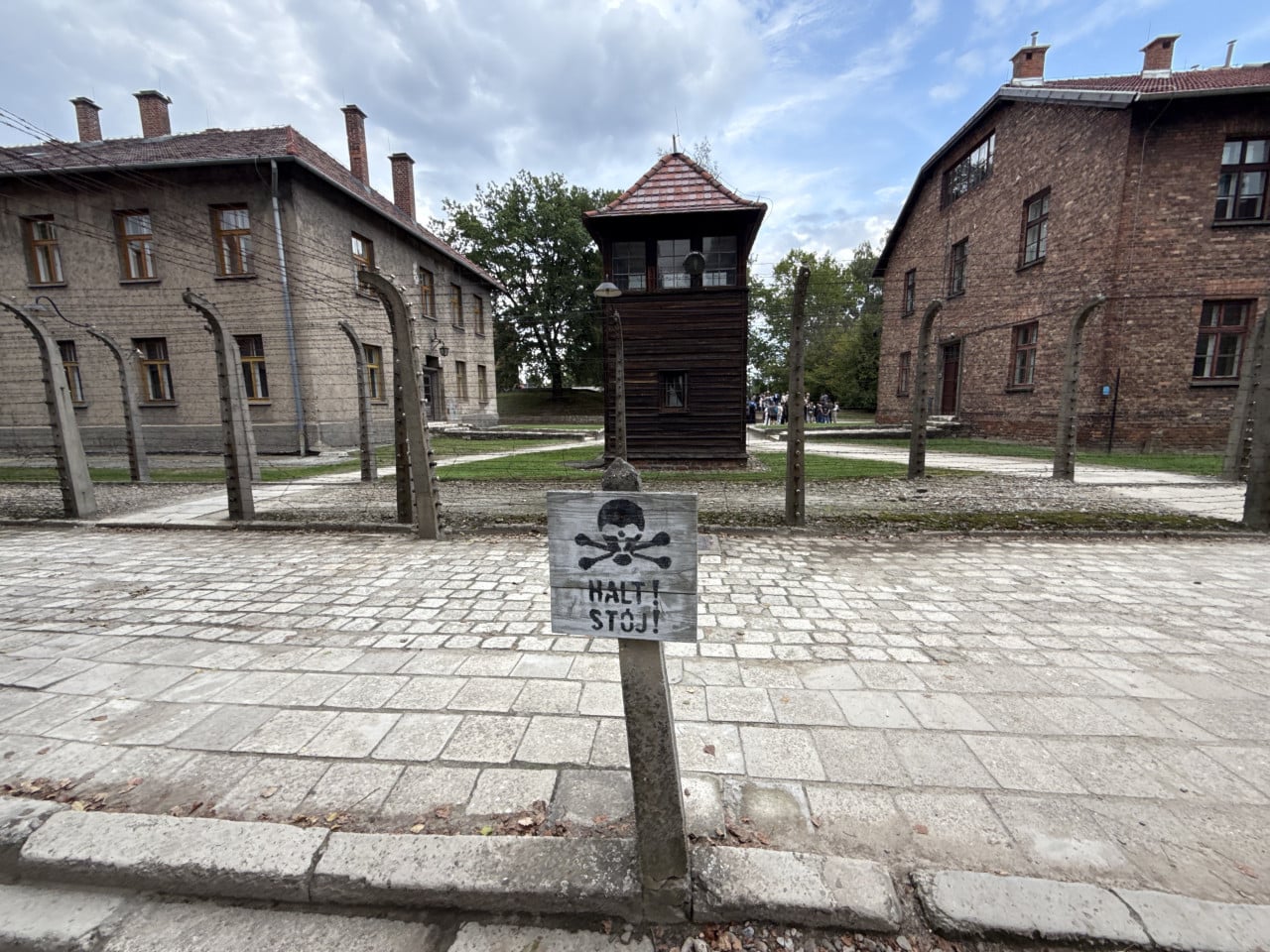 Vstup do Auschwitz s nápisem Halt! Stój!, strážní věž a cihlové budovy za plotem.