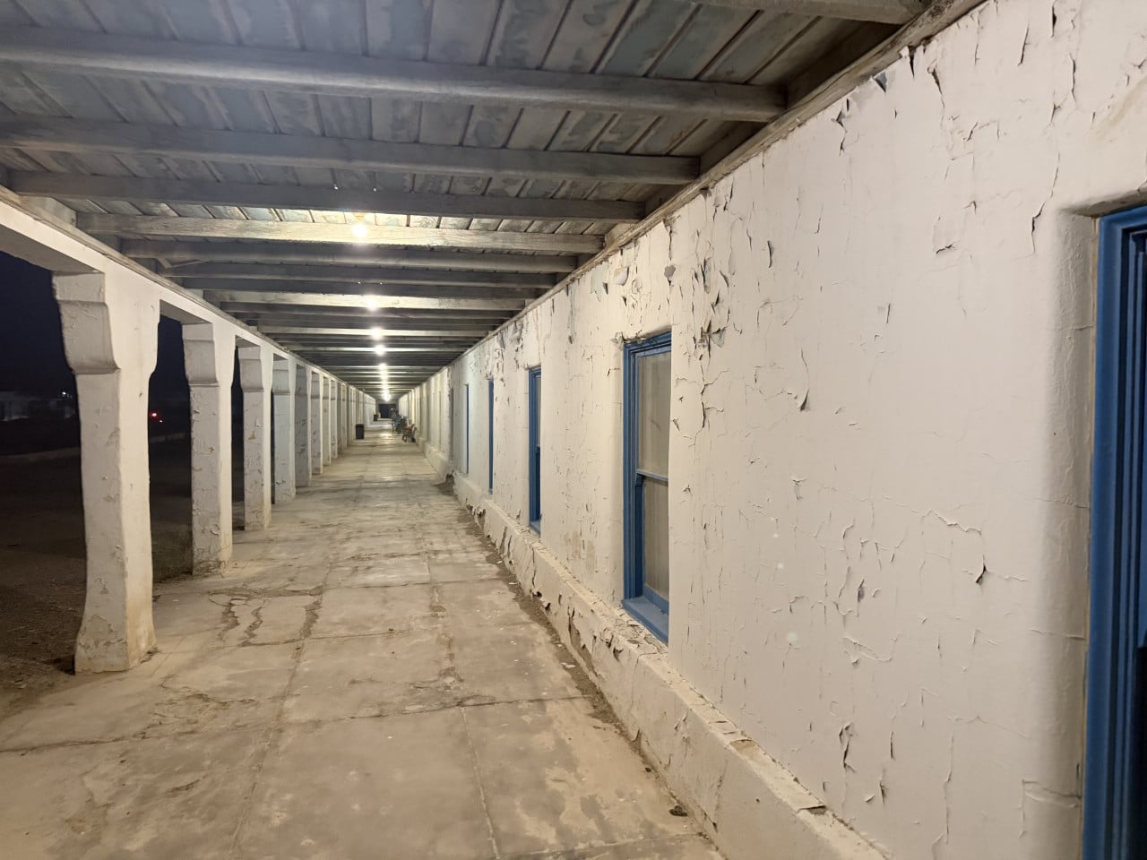 Noční chodba starého hotelu s oprýskanými bílými stěnami a modrými okny.