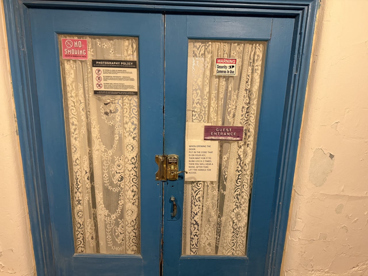 Modré hotelové dveře s krajkovými záclonami a cedulemi No Smoking, Photography Policy, Security Cameras In Use.