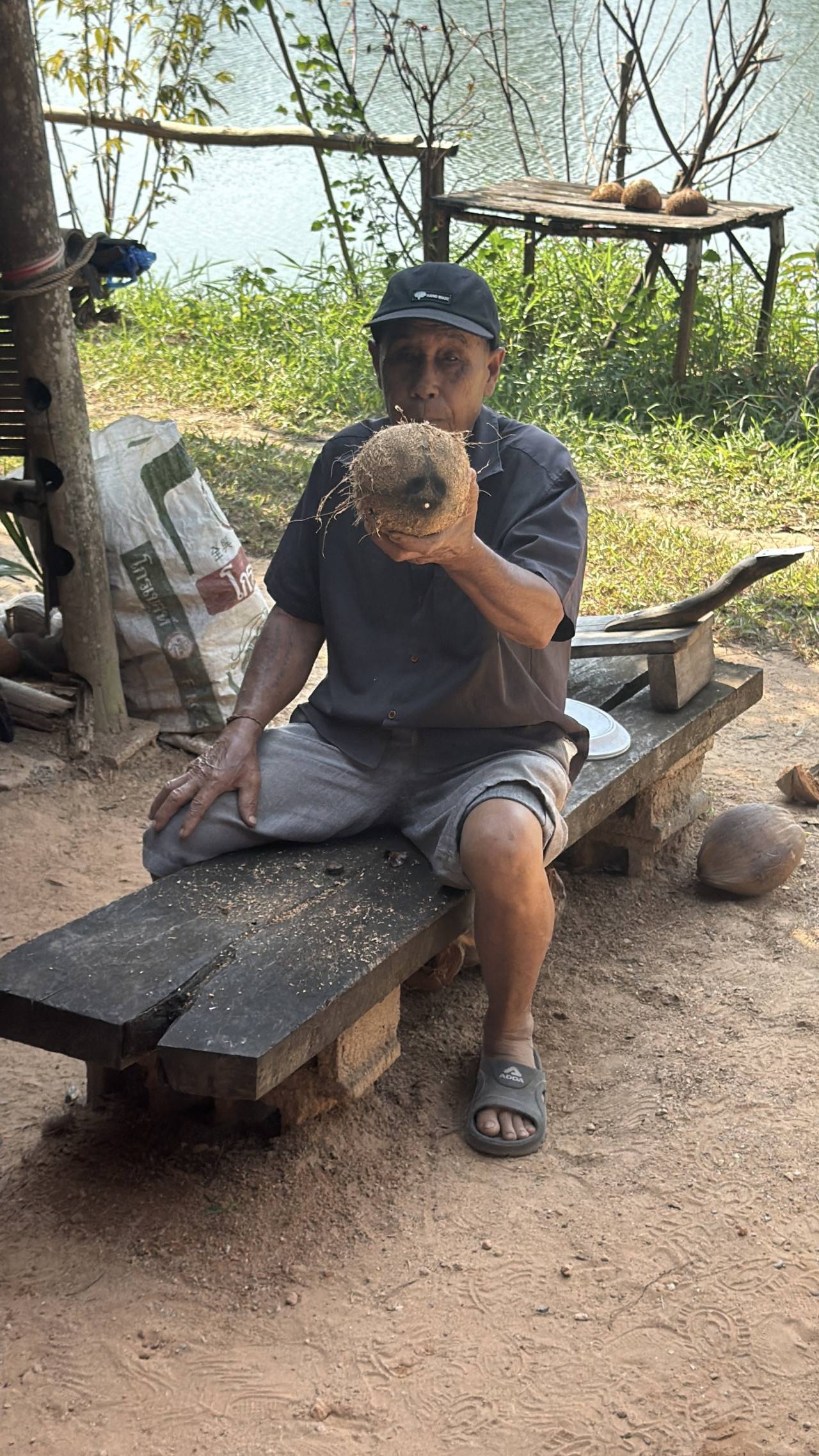 Muž sedí na lavici u řeky a ukazuje rozseknutý kokos.