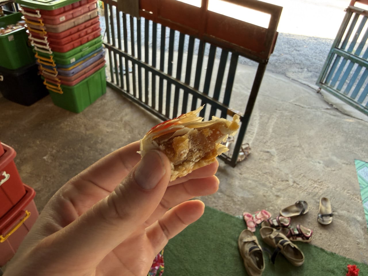 Ruka drží nakousnutý thajský koláček, v pozadí boty a plastové přepravky.