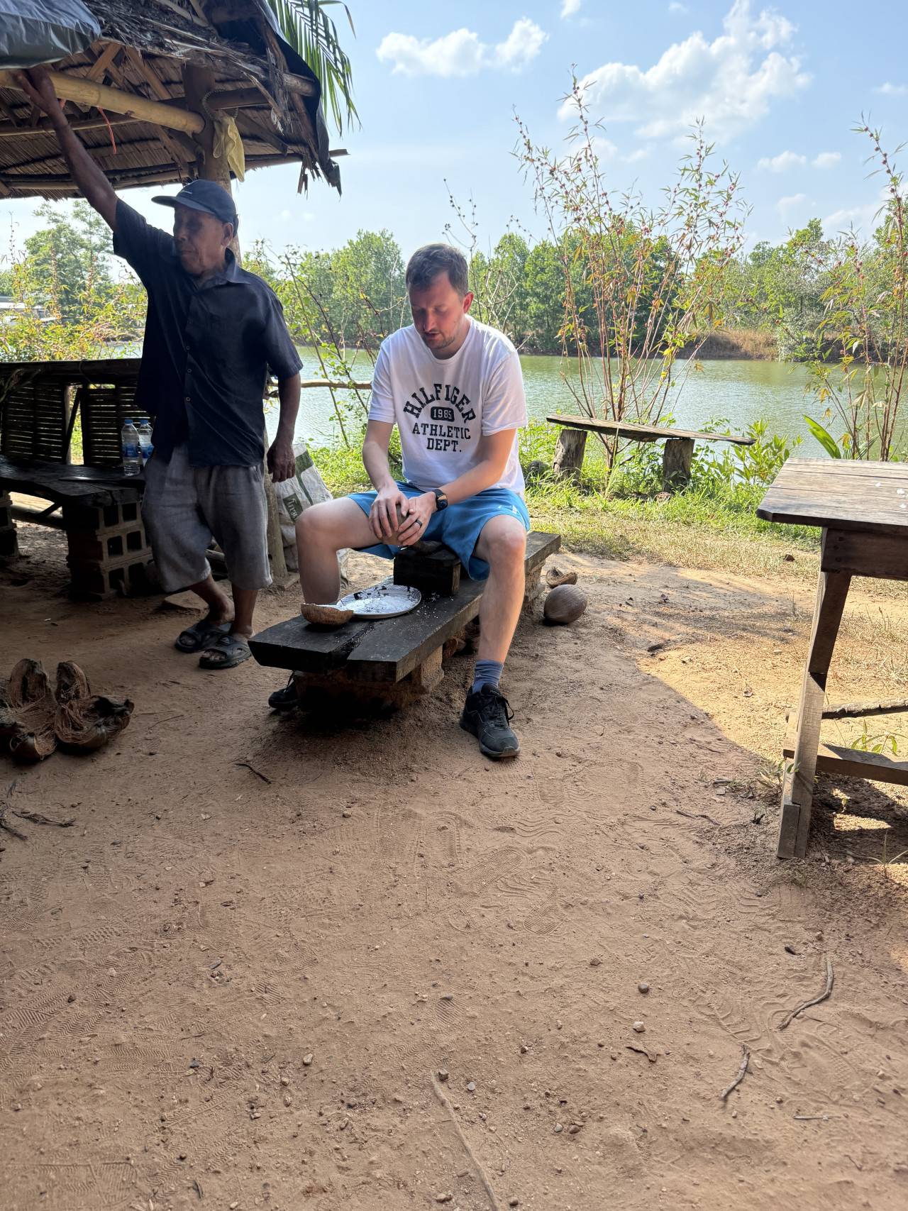 Muž v tričku HILFIGER ATHLETIC DEPT. sedí u řeky, místní muž stojí