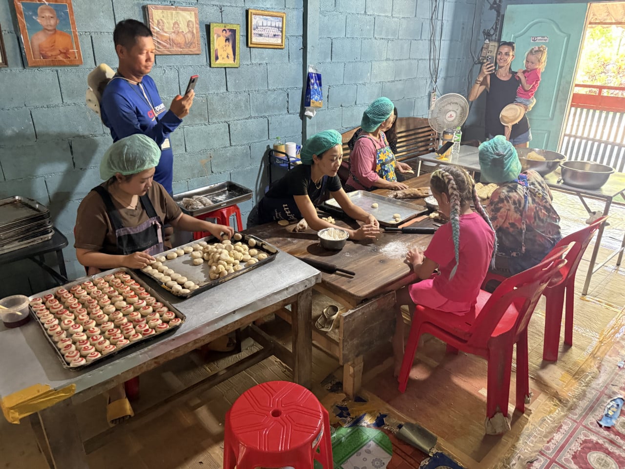 Ženy v síťkách připravují koláčky na plechách, turisté je fotí v dílně.
