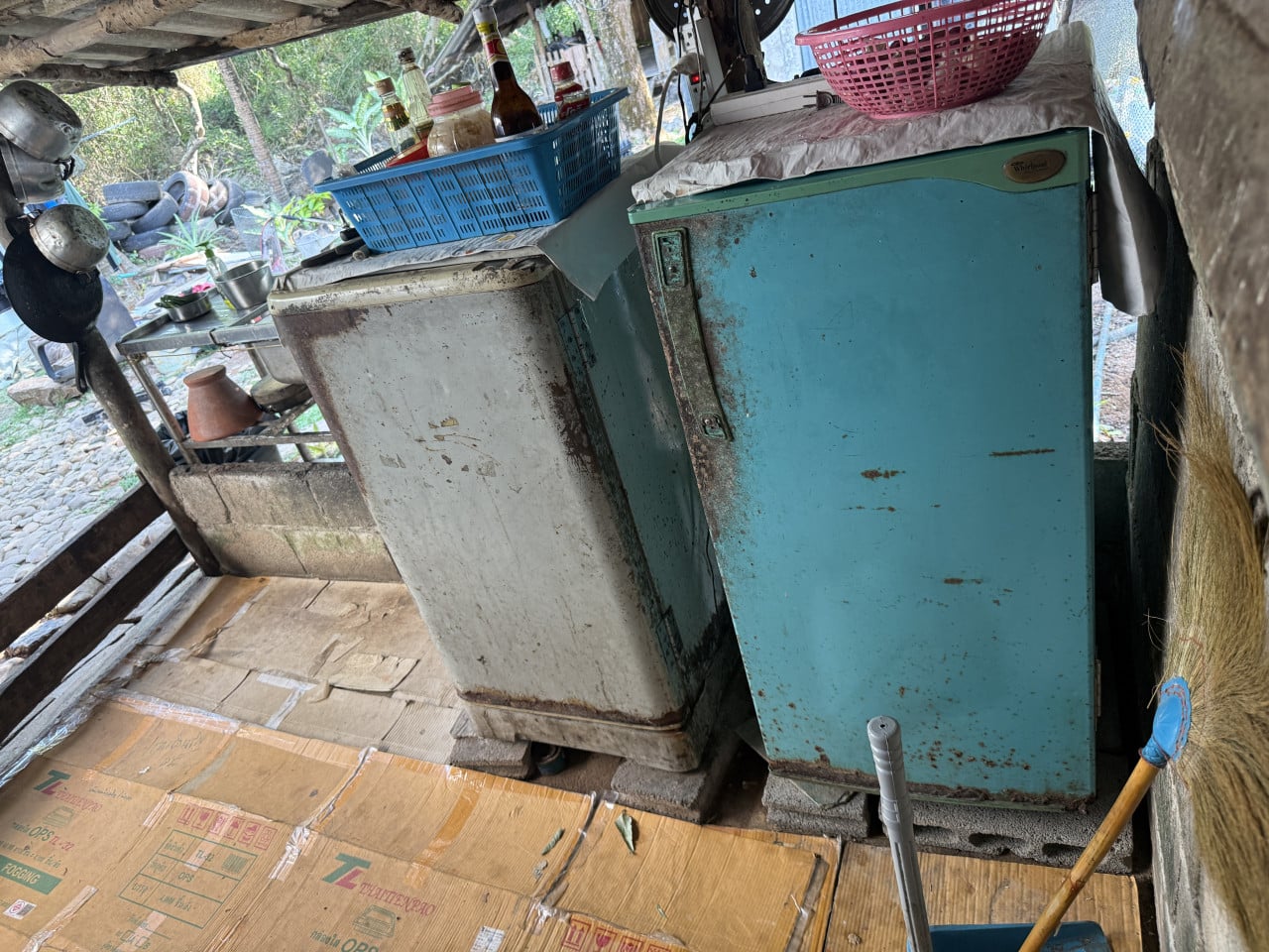 Dvě rezavé lednice, modrá s logem Whirlpool, nahoře košíky a lahve