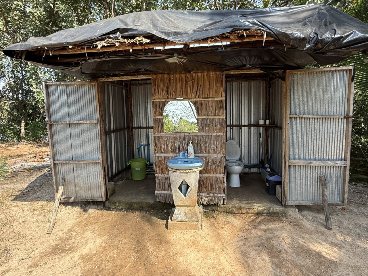 Rustikální venkovní toaleta s umyvadlem, otevřené plechové dveře, WC uvnitř.