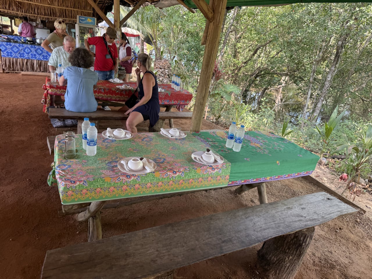 Turisté u prostřených stolů pod doškovou střechou, řeka a džungle vedle