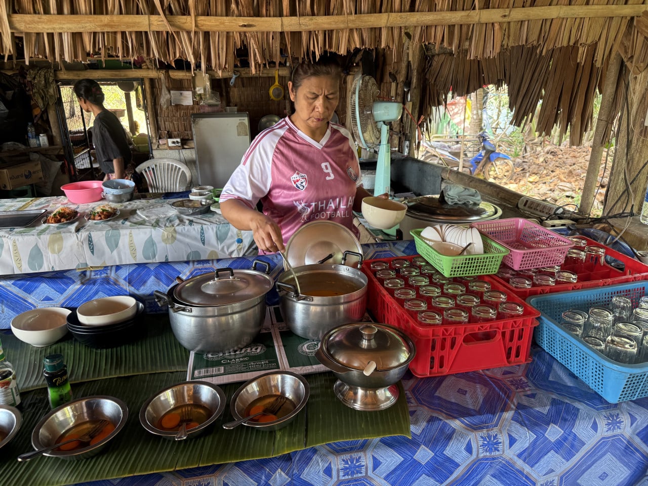 Žena v dresu THAILAND FOOTBALL 9 nabírá polévku z hrnce v přístřešku.
