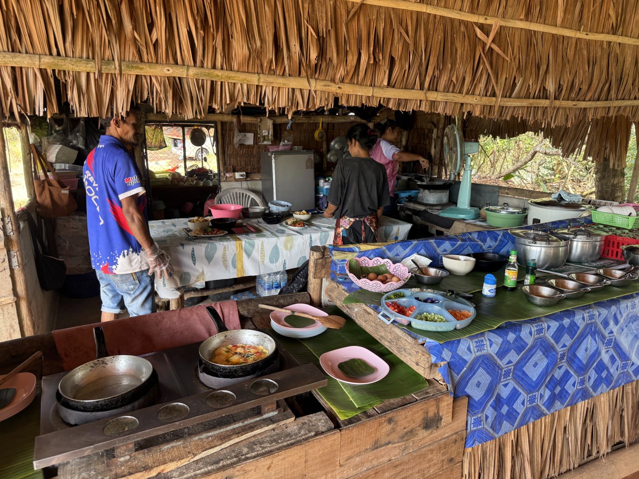 Rustikální kuchyně pod doškovou střechou, místní vaří, stoly s ingrediencemi a hrnci