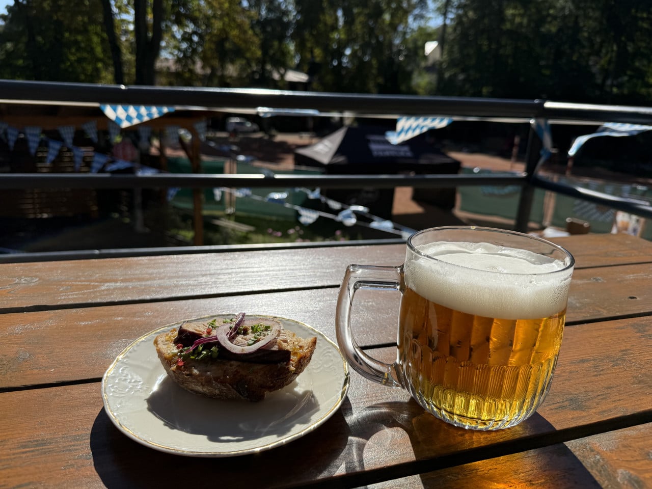 Sklenice piva a malé jídlo na dřevěném stole s bavorskou výzdobou venku.