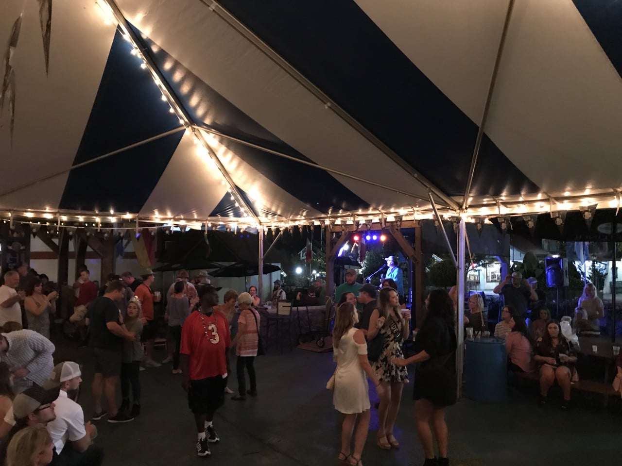 Večerní Oktoberfest pod stanem s osvětlením, lidé tančí a popíjejí u pódia