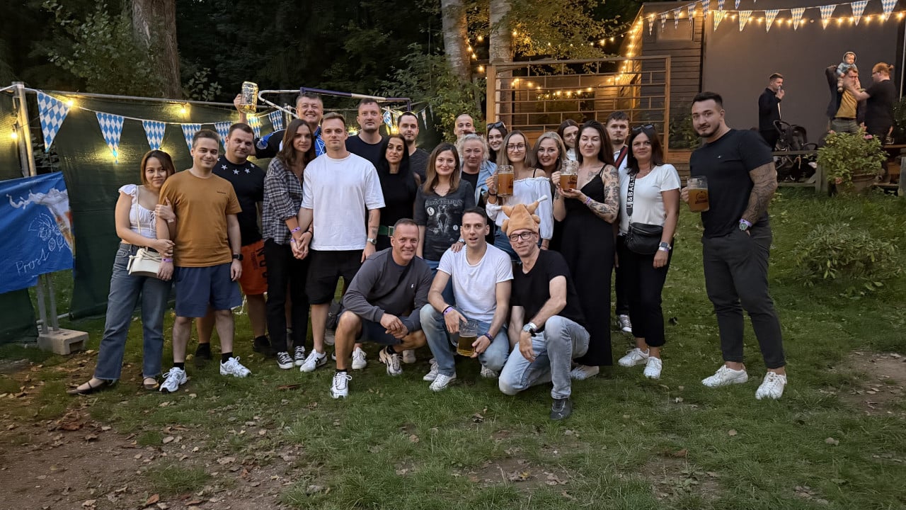 Skupina lidí s pivy pózuje na večerním Oktoberfestu pod světýlky a bavorské vlajky.