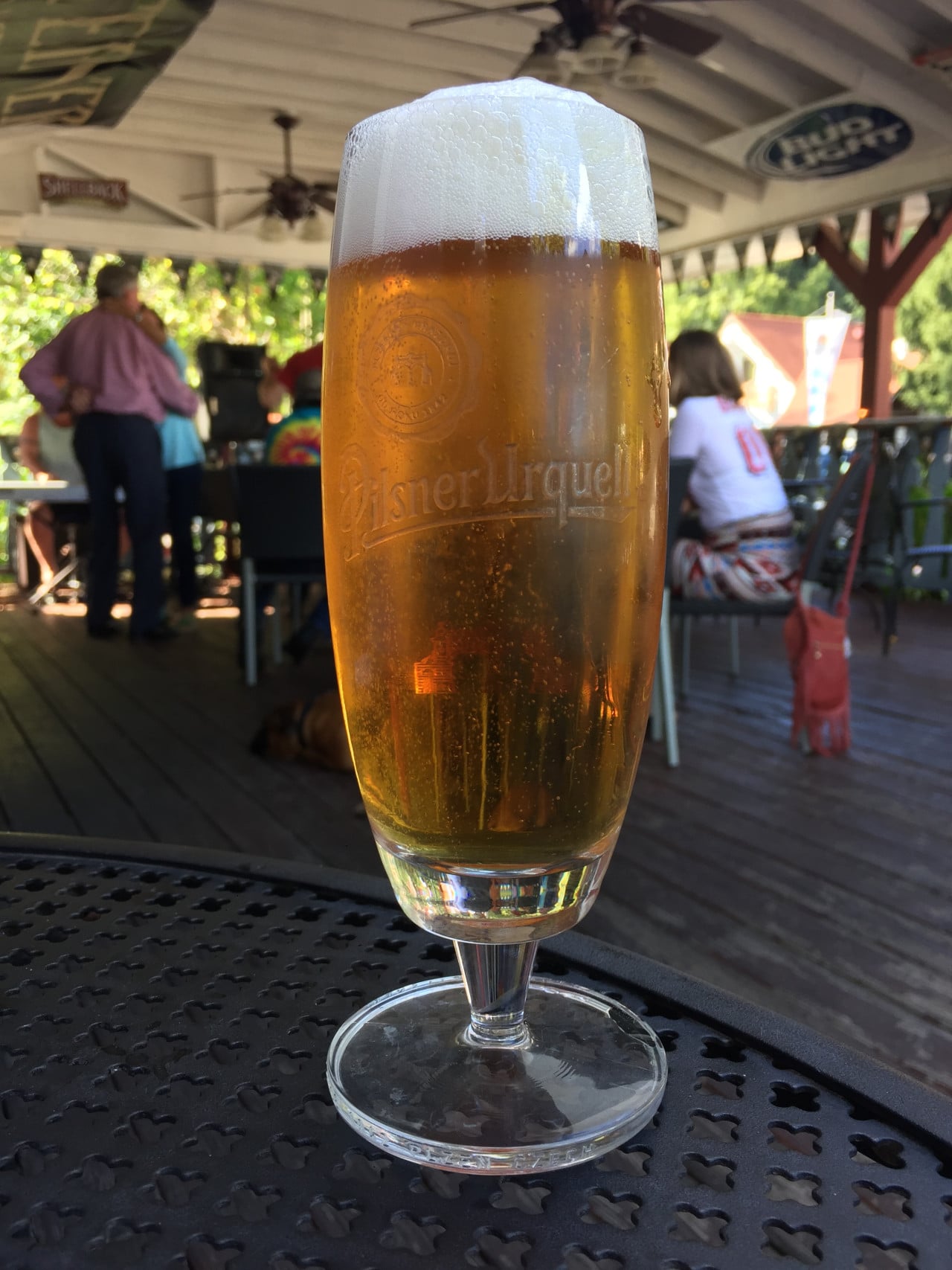 Sklenice piva na stole, v pozadí lidé na venkovní terase s logem Bud Light.
