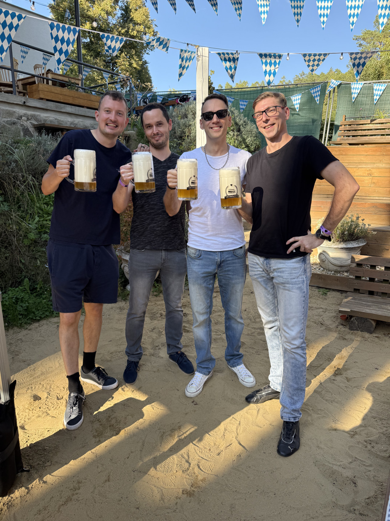 Čtyři muži na Oktoberfestu drží velké pivní korbely pod modrobílými praporky.