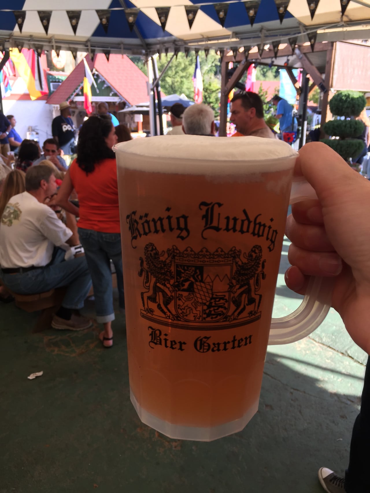 Ruka drží velký půllitr piva v biergartenu s lidmi a vlaječkami na Oktoberfestu.