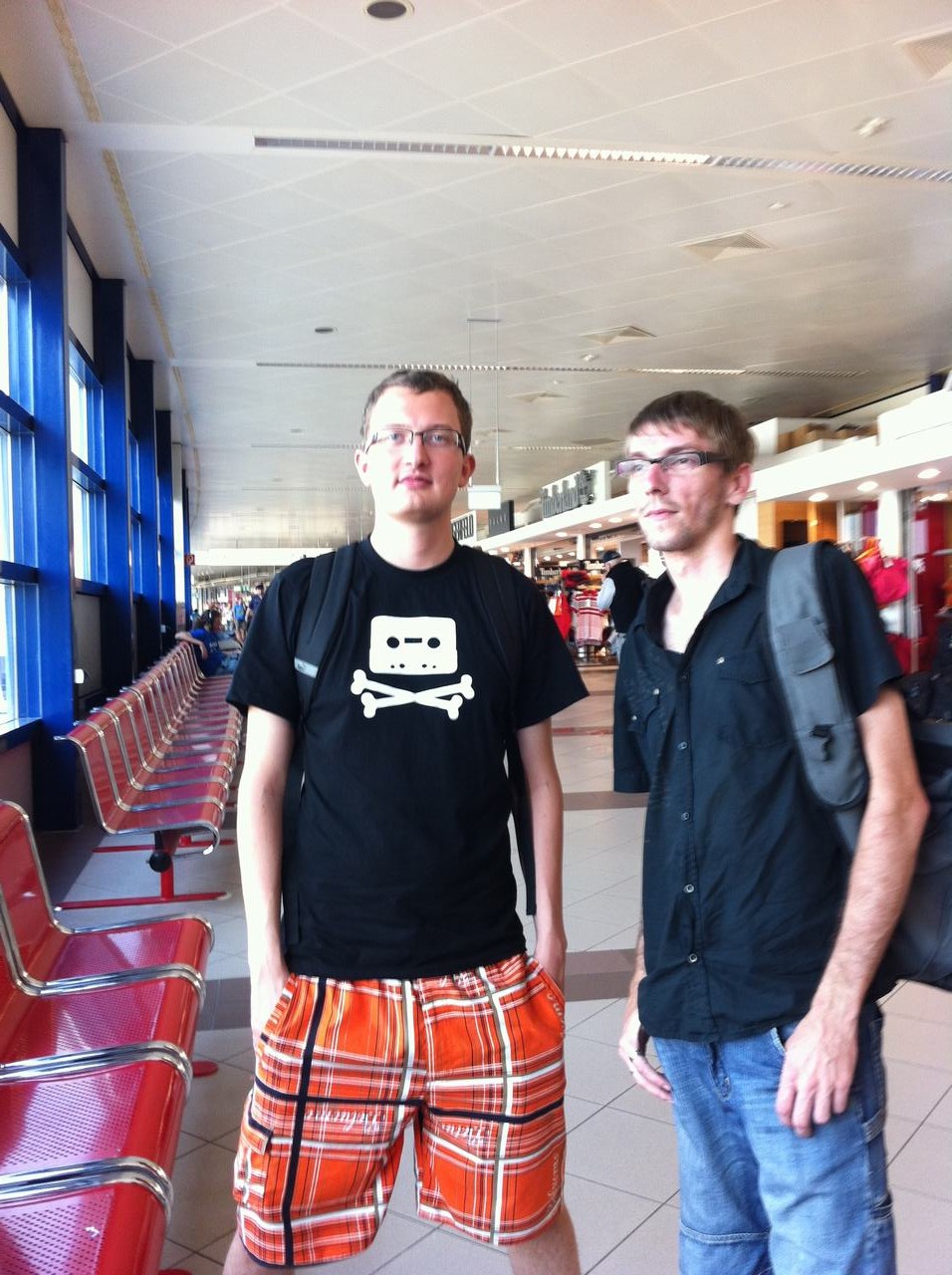 Dva muži s batohy v letištní hale, vedle červených sedaček čekárny.