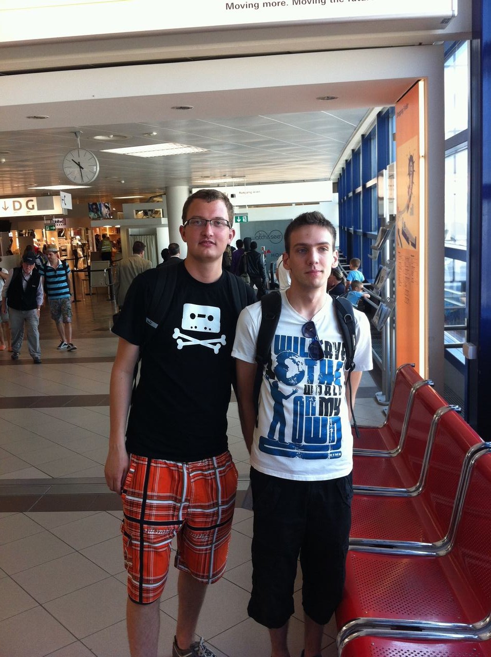 Dva mladí muži s batohy stojí na letišti u červených sedadel, pod hodinami.