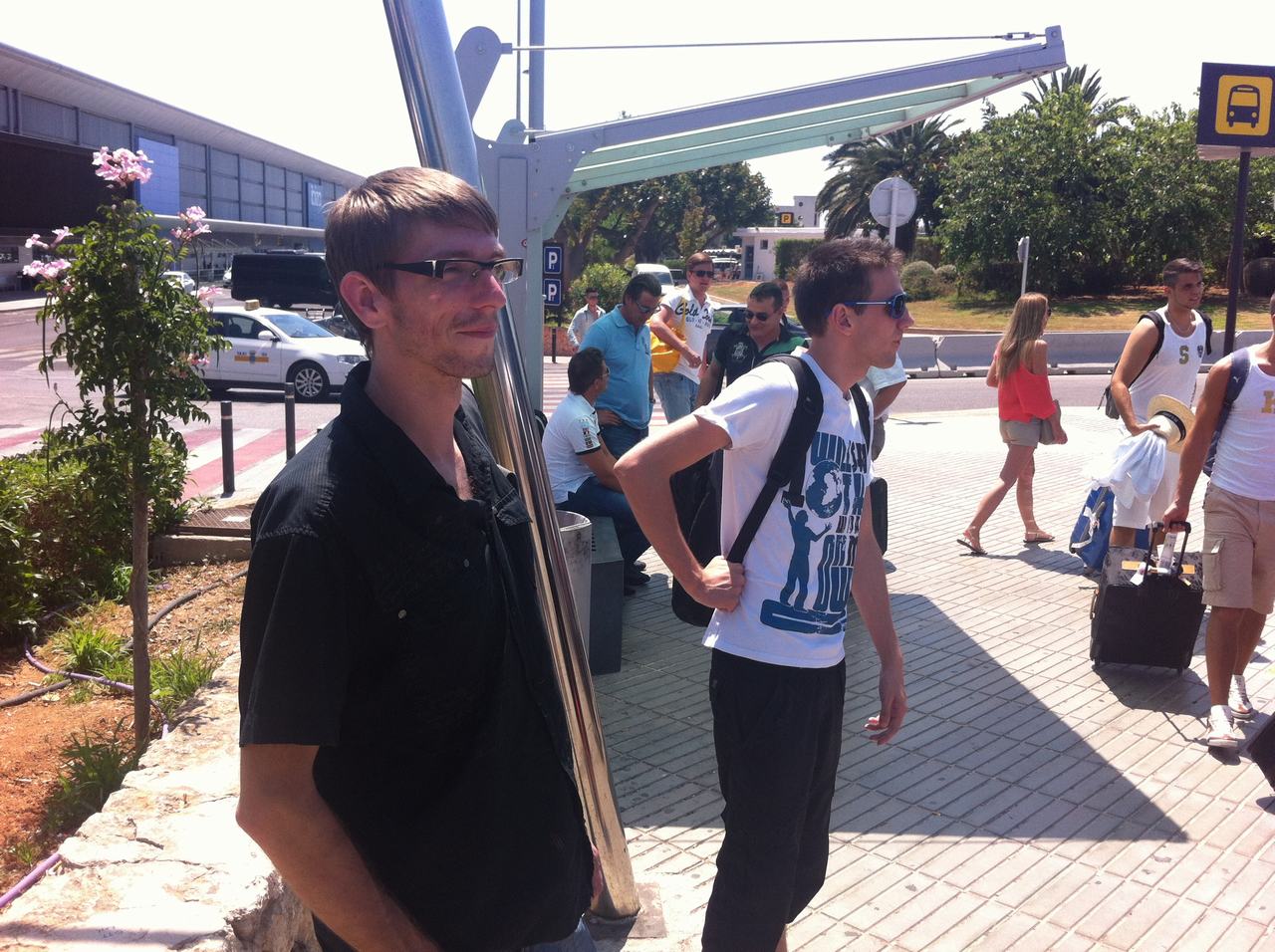 Dva muži s batohy čekají u zastávky před letištním terminálem, cestující s kufry.