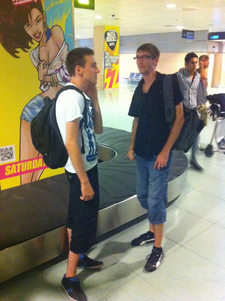 Dva muži s batohy u letištního pásu, vedle plakátu SATURDA.