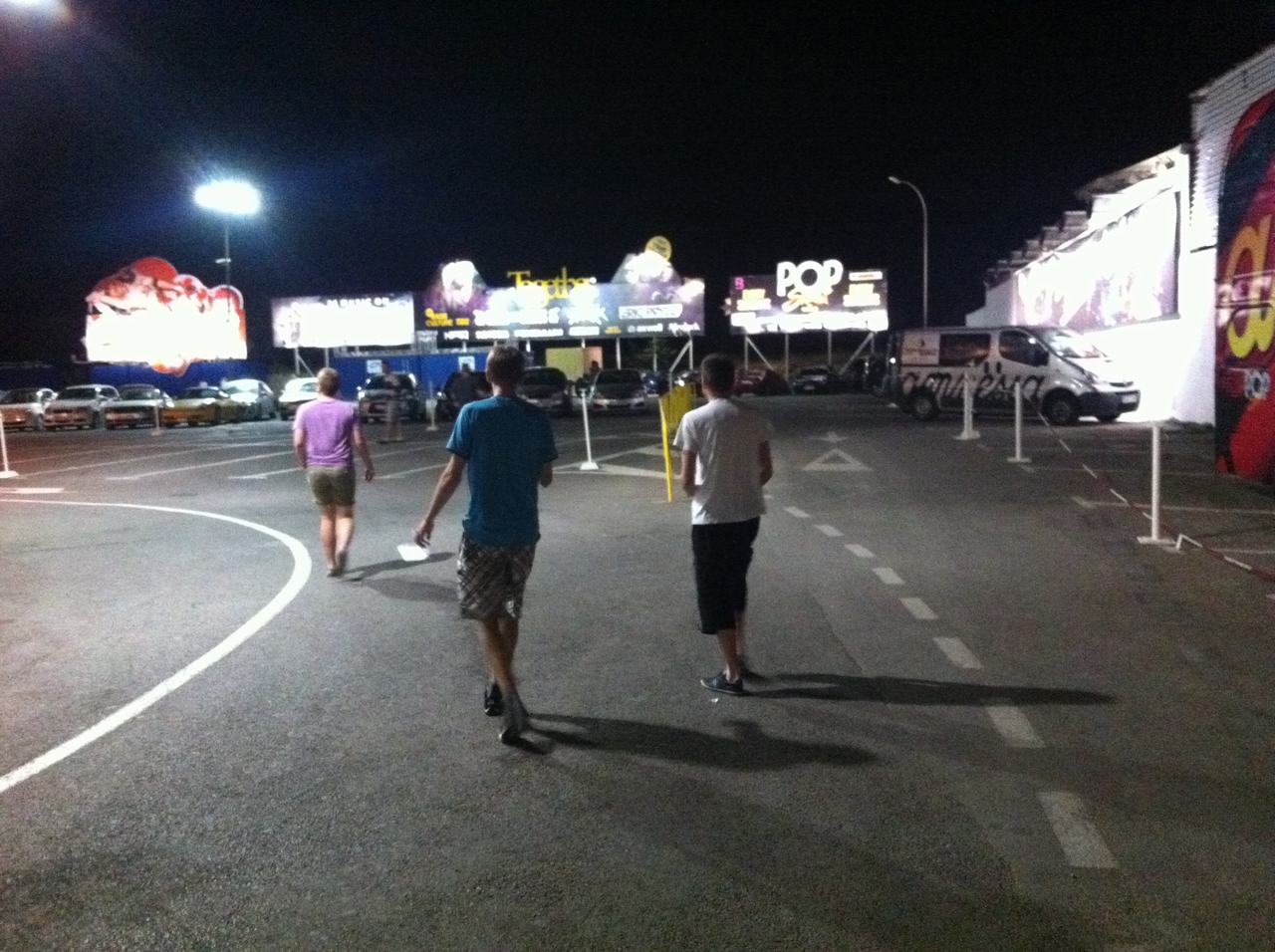 Tři muži jdou v noci parkovištěm k billboardům s nápisem POP.