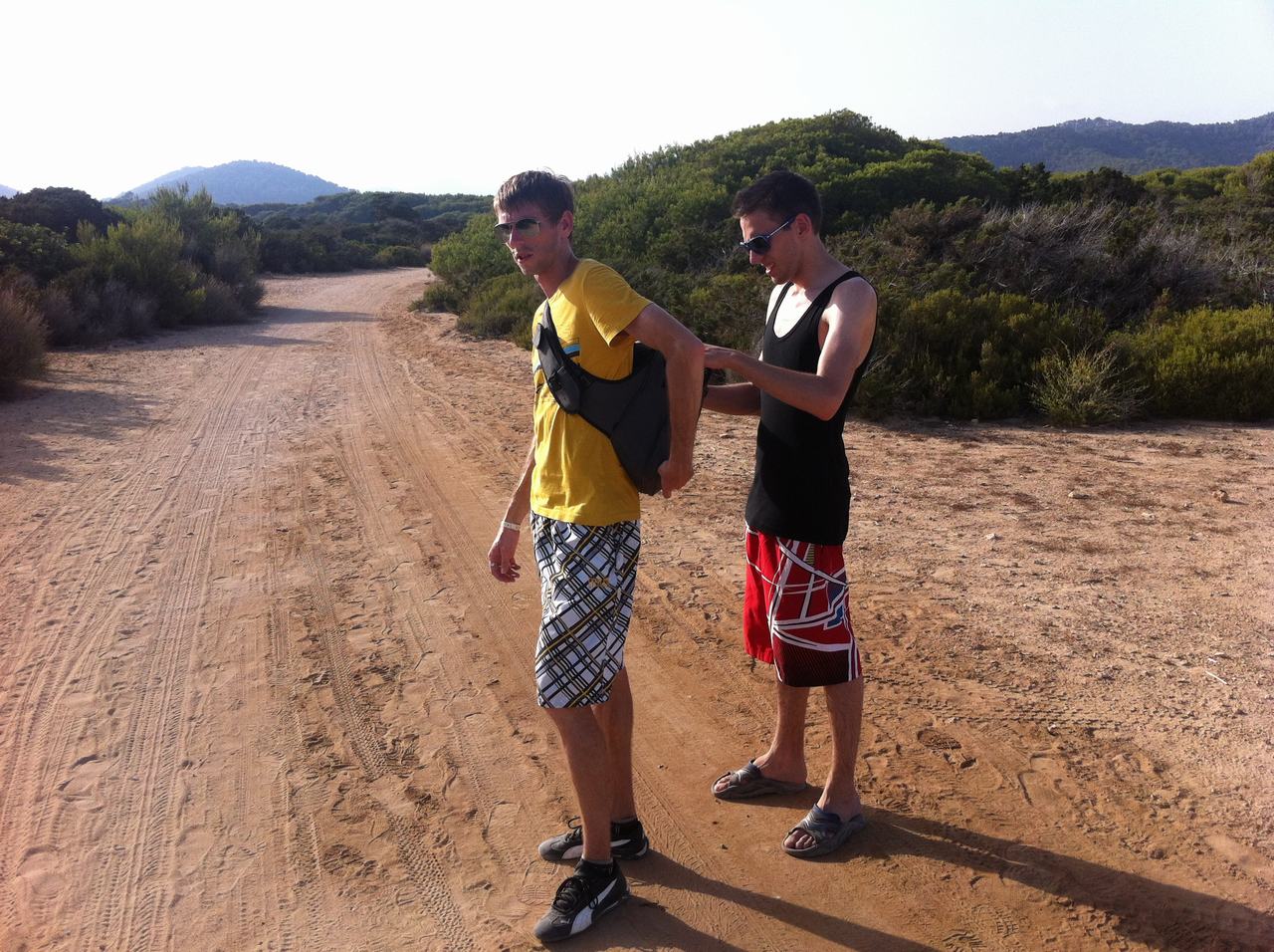 Dva muži v kraťasech na písčité cestě, jeden upravuje druhému vestu.