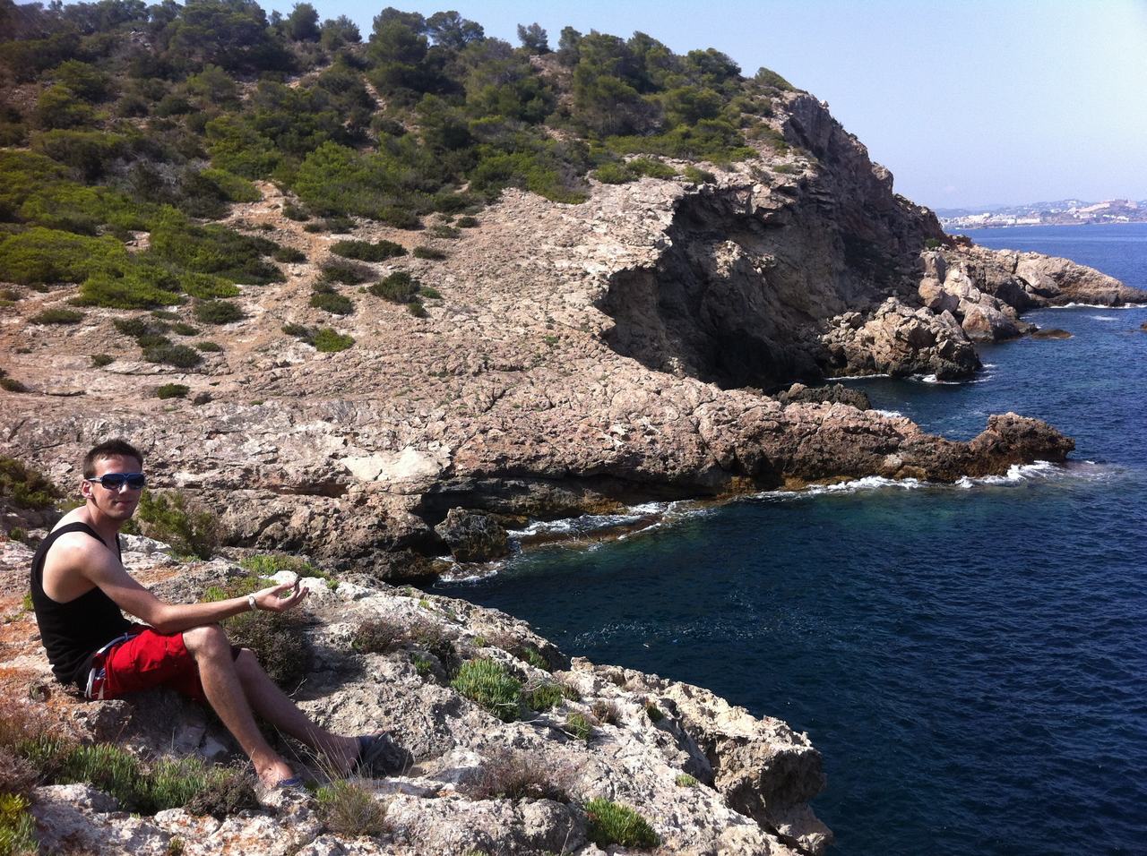 Muž sedí na skalnatém útesu nad mořem, v pozadí zalesněný svah