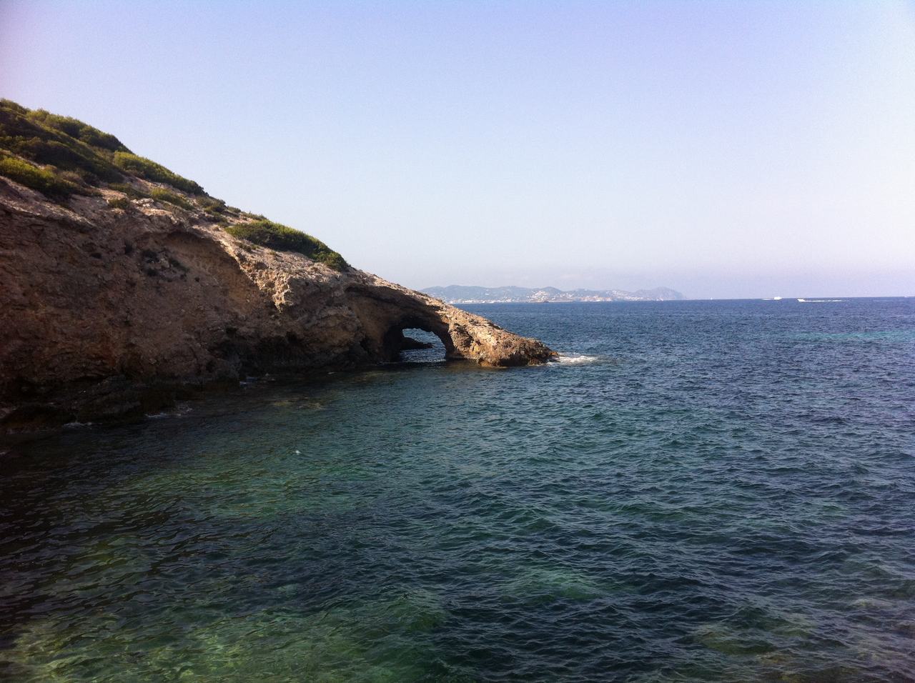 Skalnatý výběžek s přírodním obloukem nad mořem, v dálce kopce ostrova