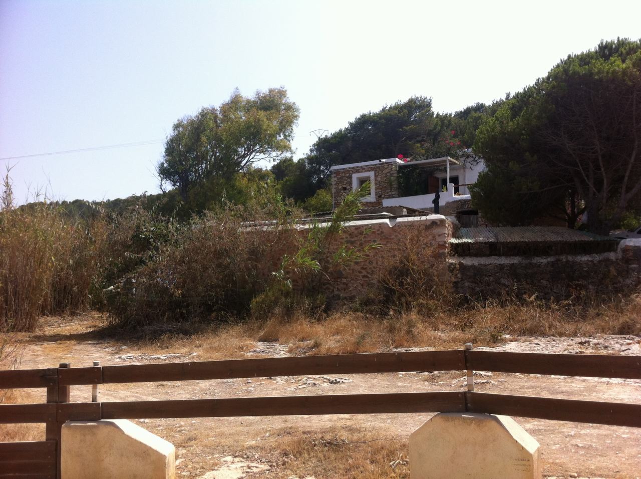 Kamenný dům s bílou terasou uprostřed stromů, suchá vegetace a plot v popředí