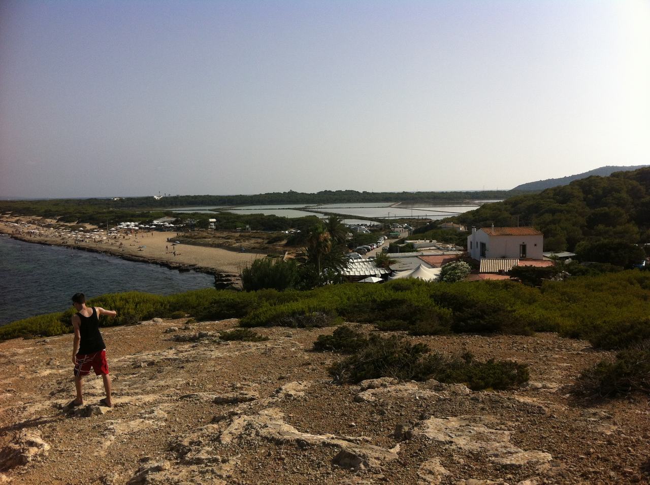Muž na skalnatém svahu hledí na pláž, zátoku a domy na Ibize