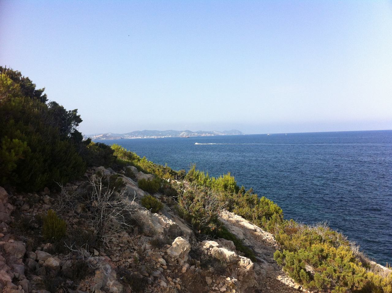 Skalnaté pobřeží s křovinami, výhled na moře a vzdálené pobřeží