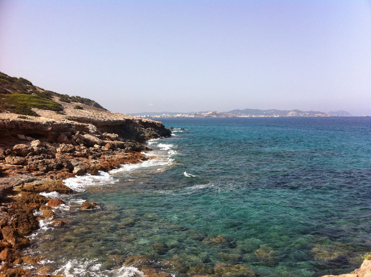 Skalnaté pobřeží s průzračně tyrkysovým mořem, v dálce město na obzoru