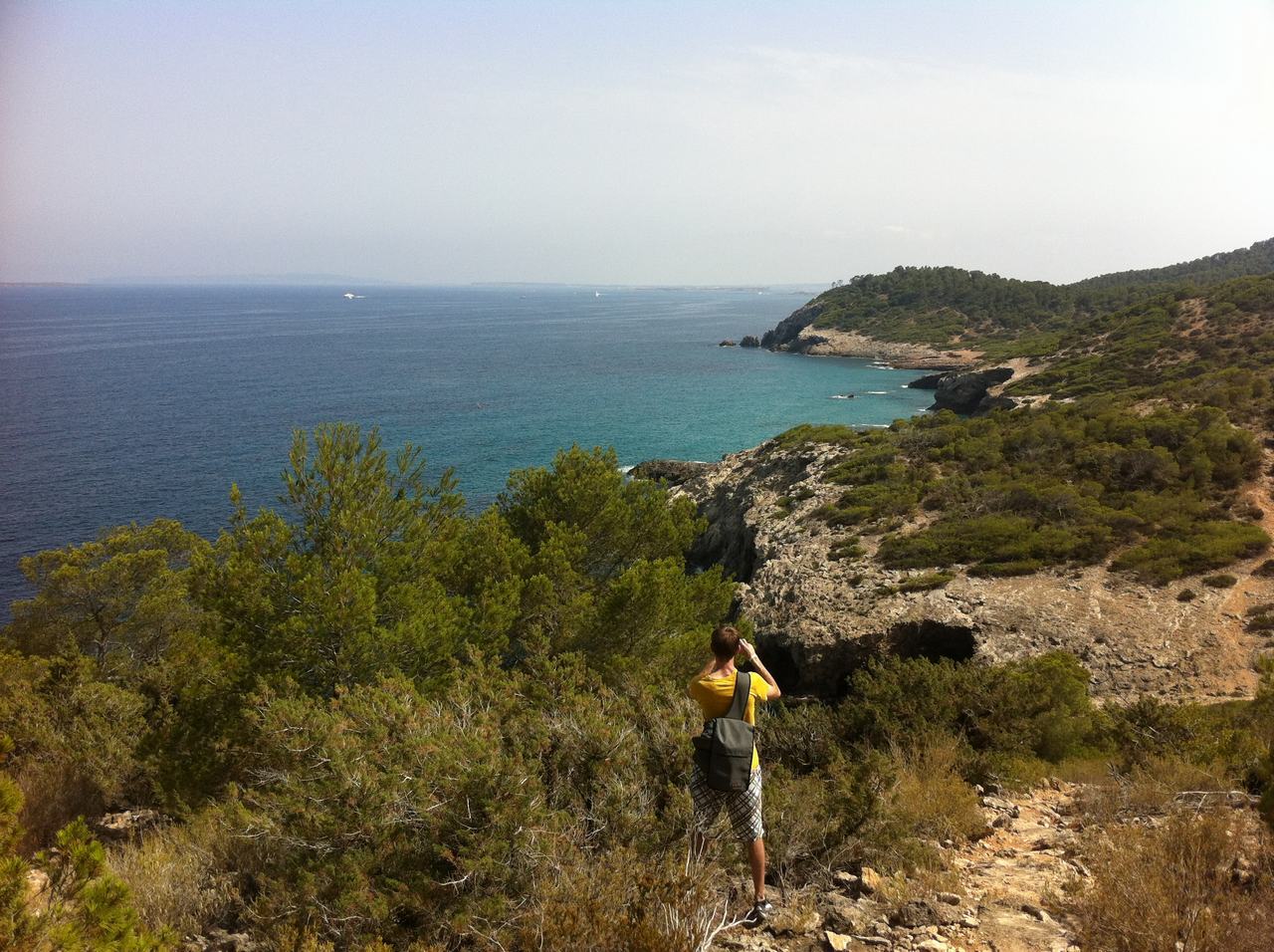 Muž s batohem fotí tyrkysové moře z skalnatého pobřeží Ibizy.
