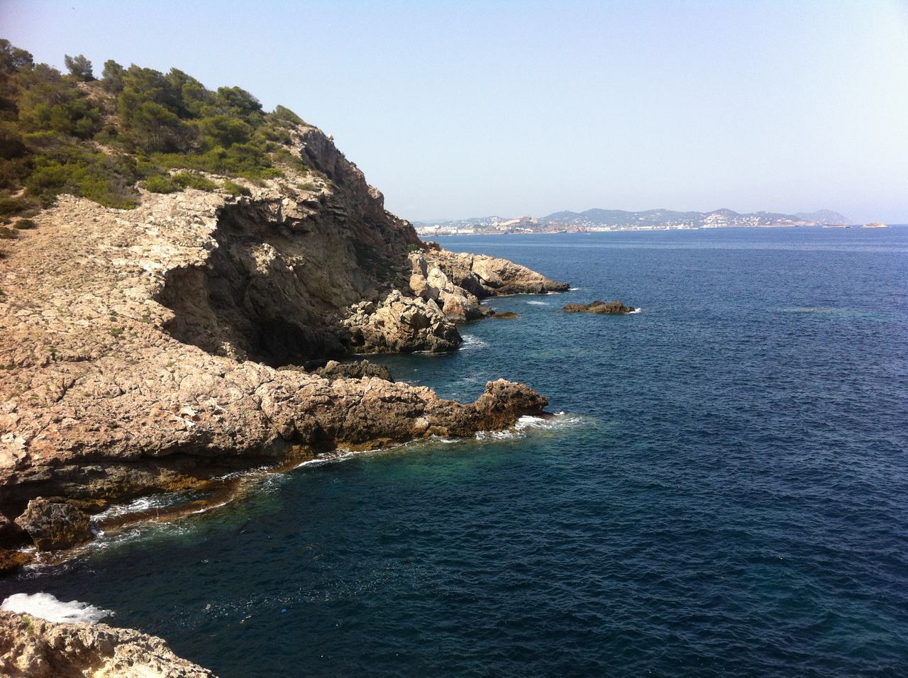 Skalnaté pobřeží s borovicemi nad modrým mořem, v dálce ostrovní panorama.