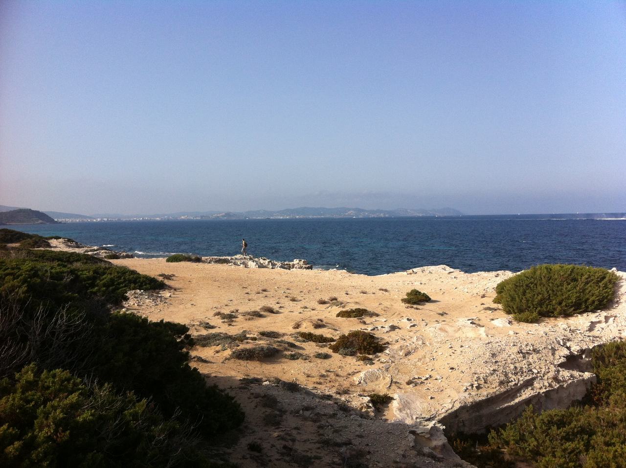 Skalnaté pobřeží s křovinami, písčitá cesta a moře, osamělá postava v dálce