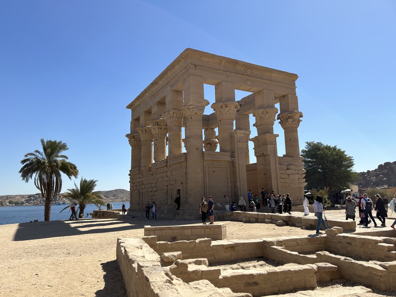 Chrám Philae u vody, obklopený palmami a turisty pod modrou oblohou