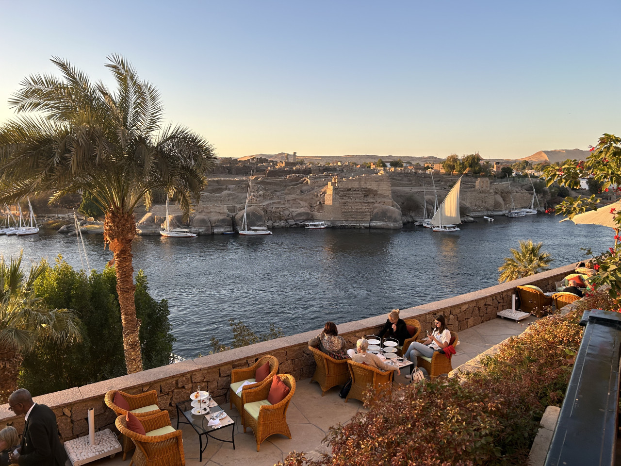 Terasa hotelu u Nilu s palmami, loděmi a ruinami Philae v pozadí