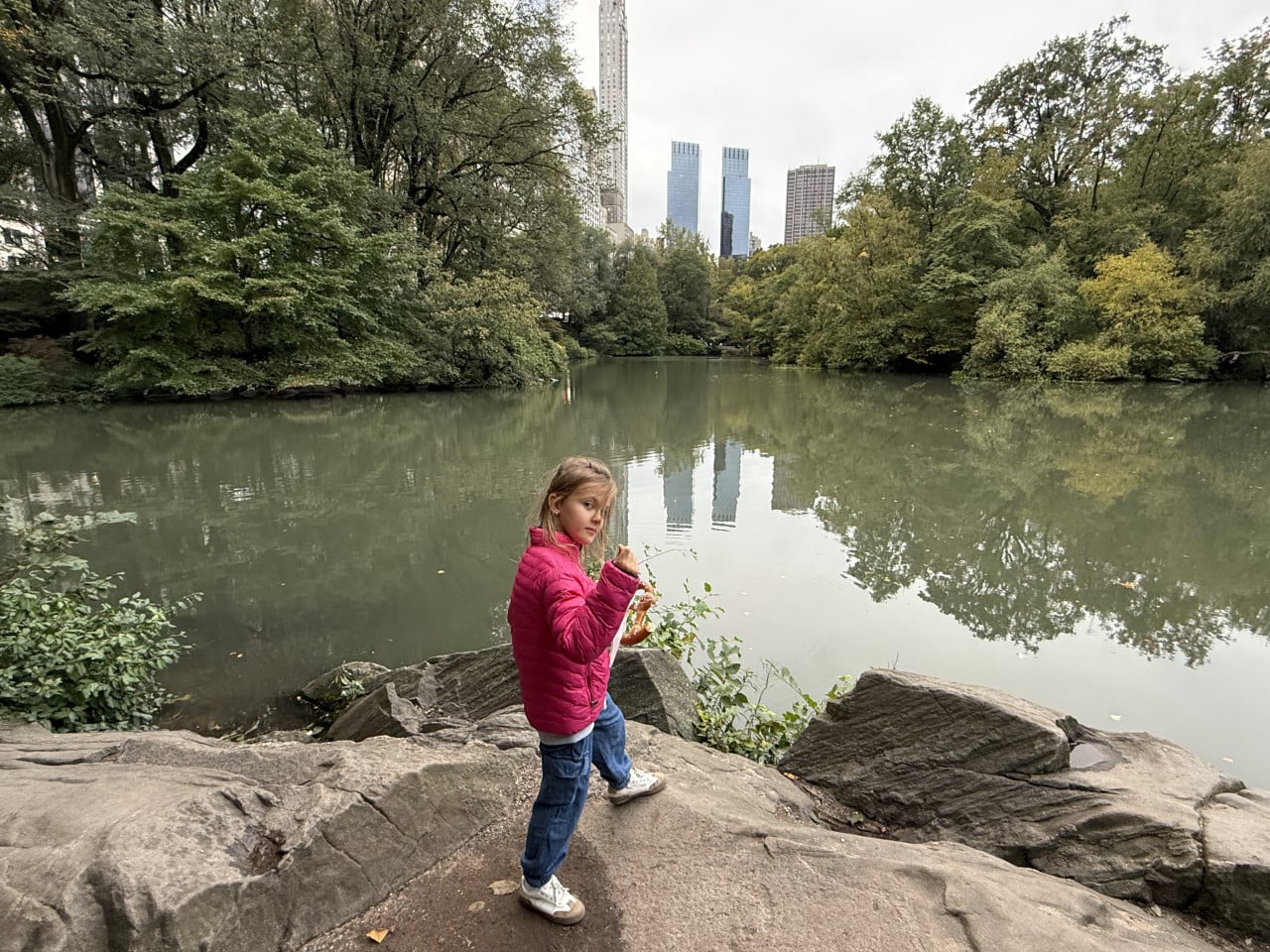 Dívka v růžové bundě stojí u jezírka v Central Parku s mrakodrapy v pozadí
