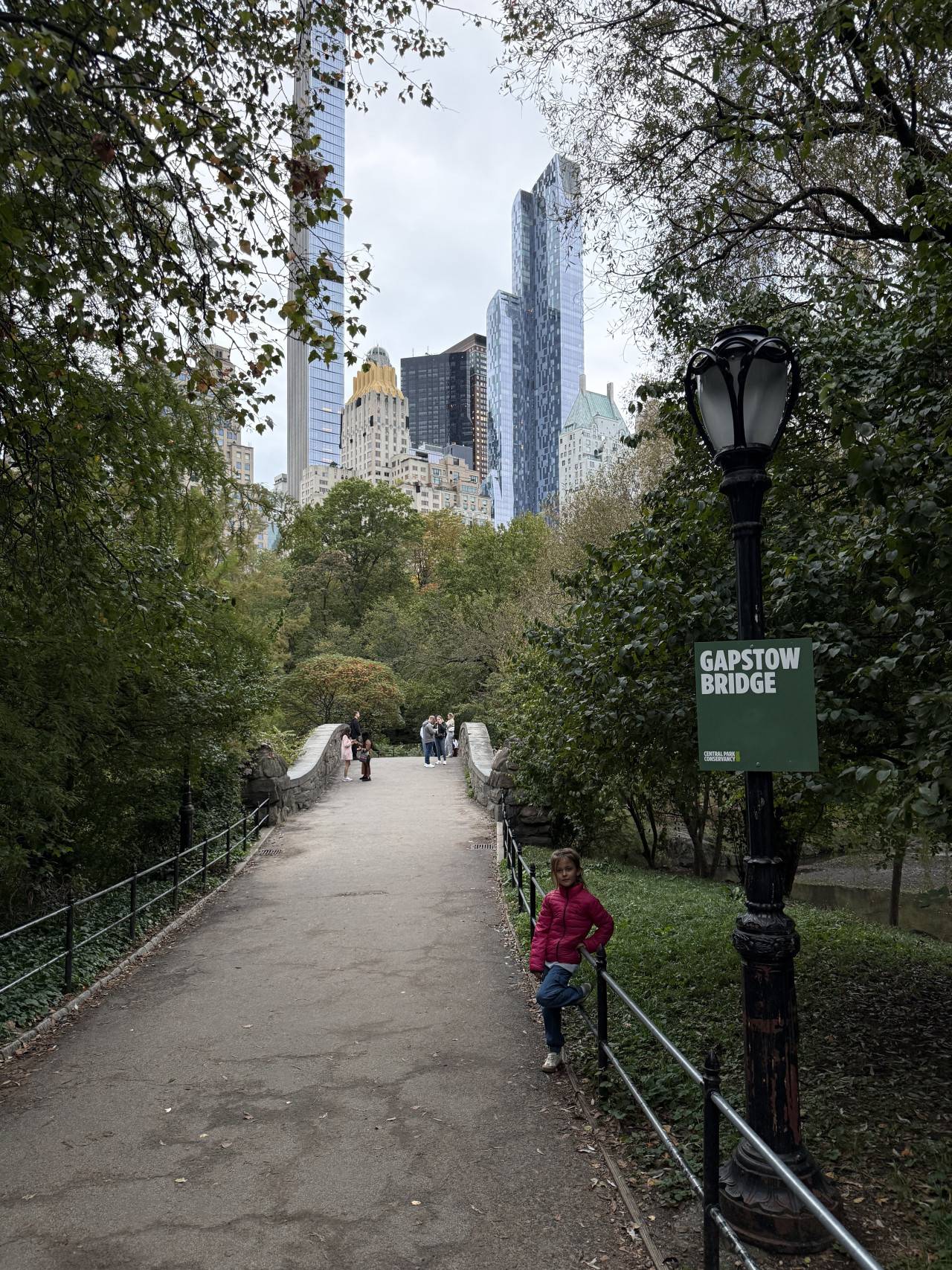 Central Park walkway by Gapstow Bridge, Manhattan skyline behind trees, child in pink jacket