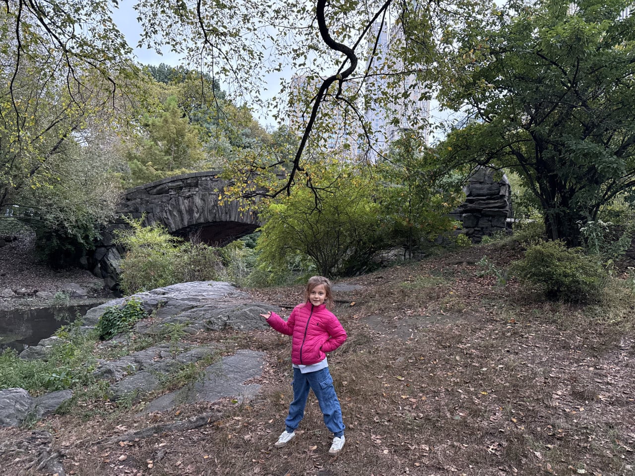Dívka v růžové bundě stojí v Central Parku u kamenného mostu.