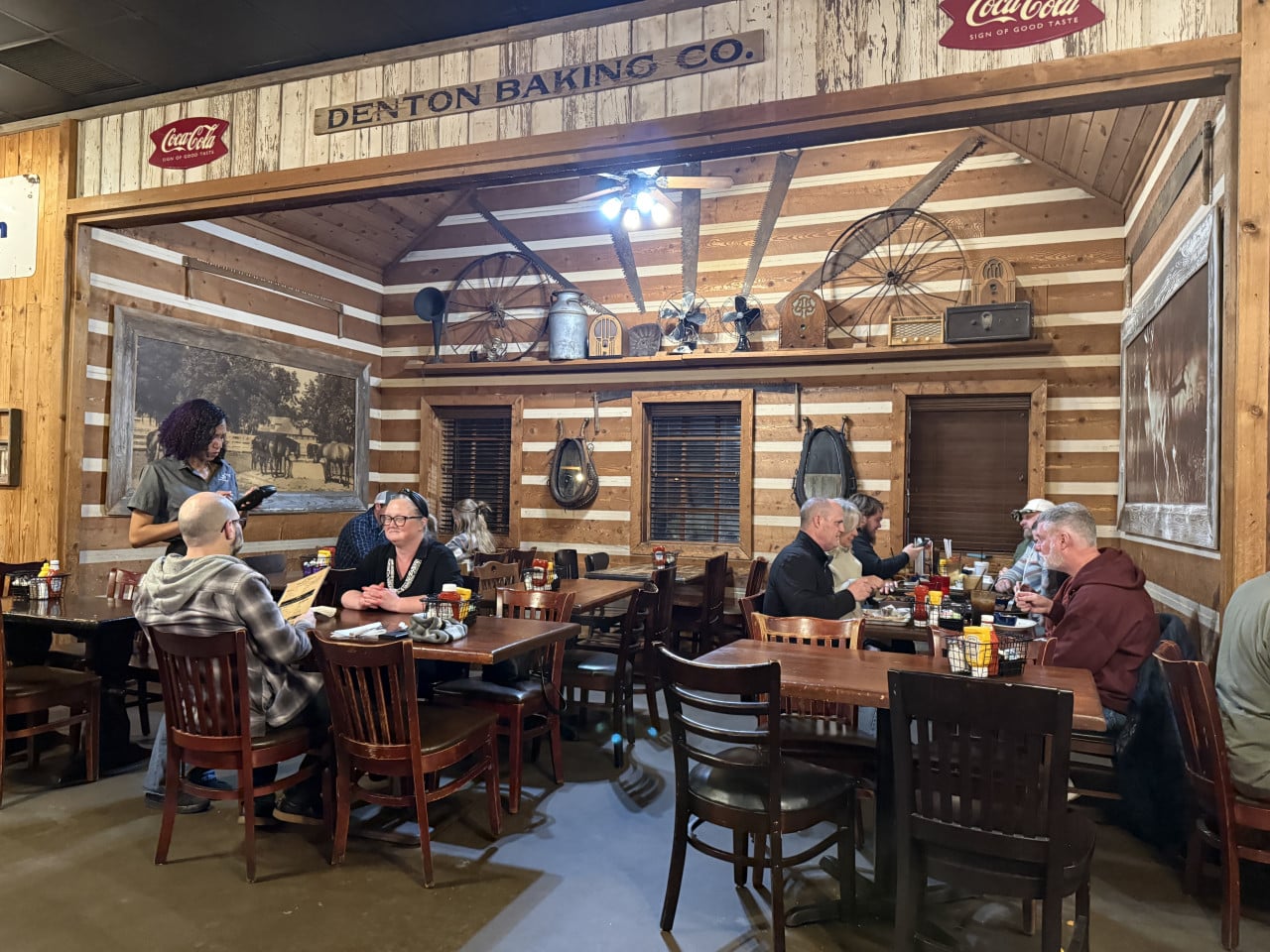 Hosté u stolů v rustikální jídelně, nápis DENTON BAKINS CO., Coca-Cola.