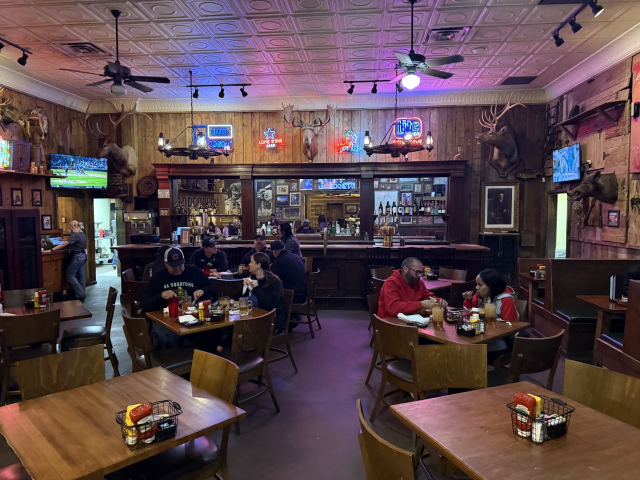 Interiér restaurace s barem, hosty, trofejemi a neonovými nápisy BUD LIGHT, LONE STAR, Lite.