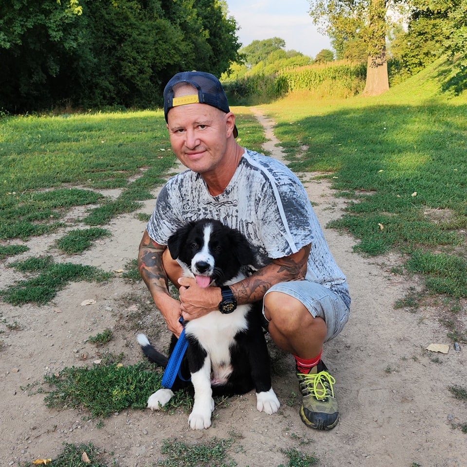 Muž v kšiltovce klečí na cestě a objímá černobílé štěně na vodítku.