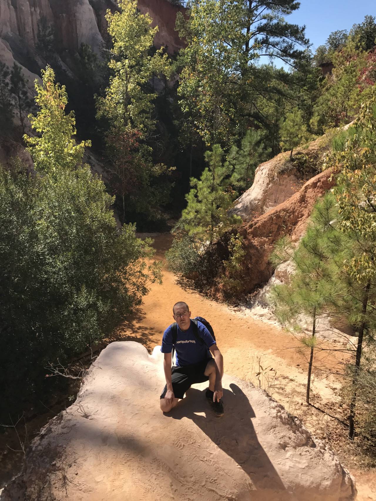 Muž klečí na písčitém útesu v Providence Canyon, obklopeném stromy a barevnými skalami