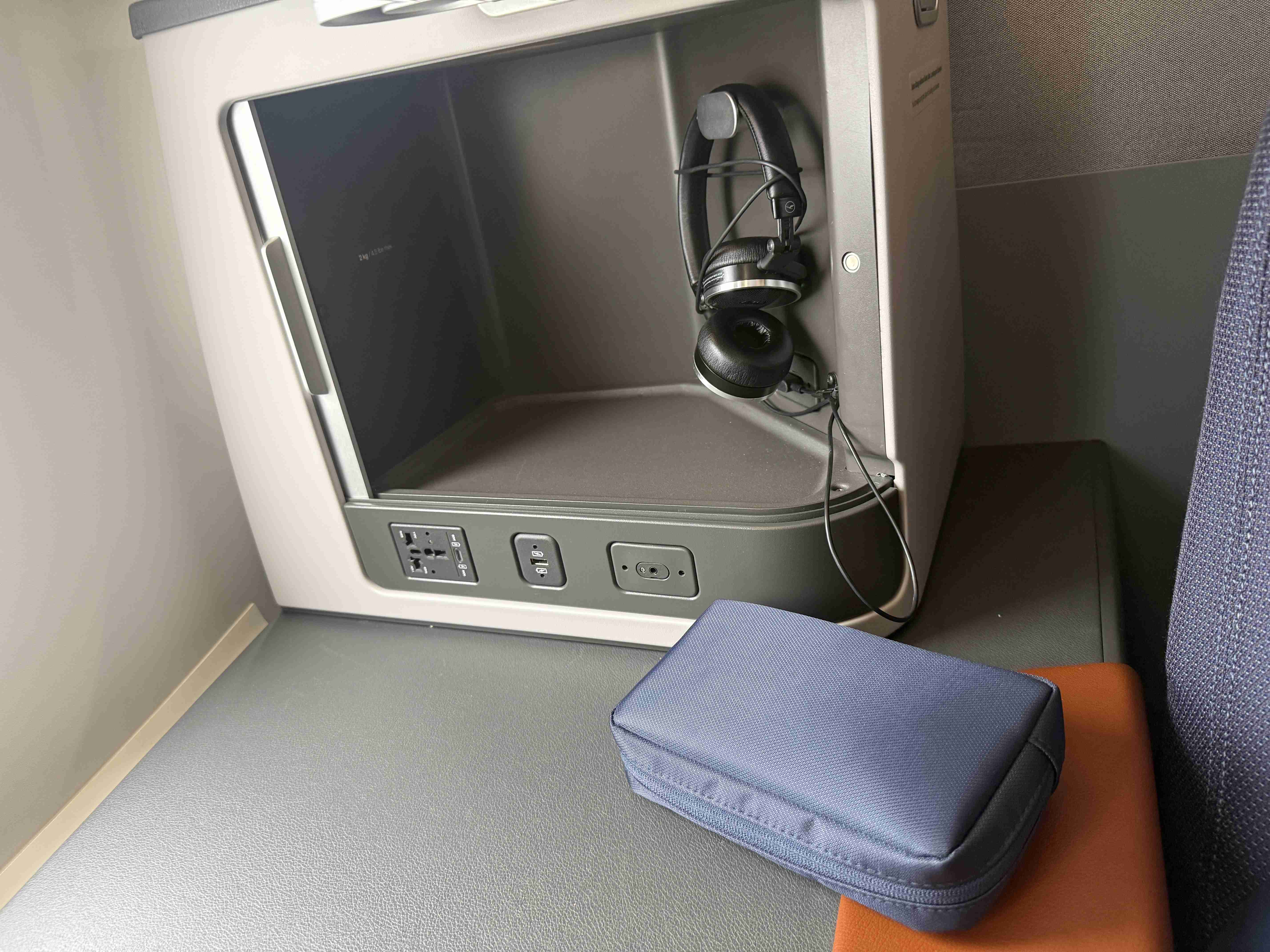 Přihrádka sedadla v letadle se sluchátky, zásuvkou a modrým pouzdrem.