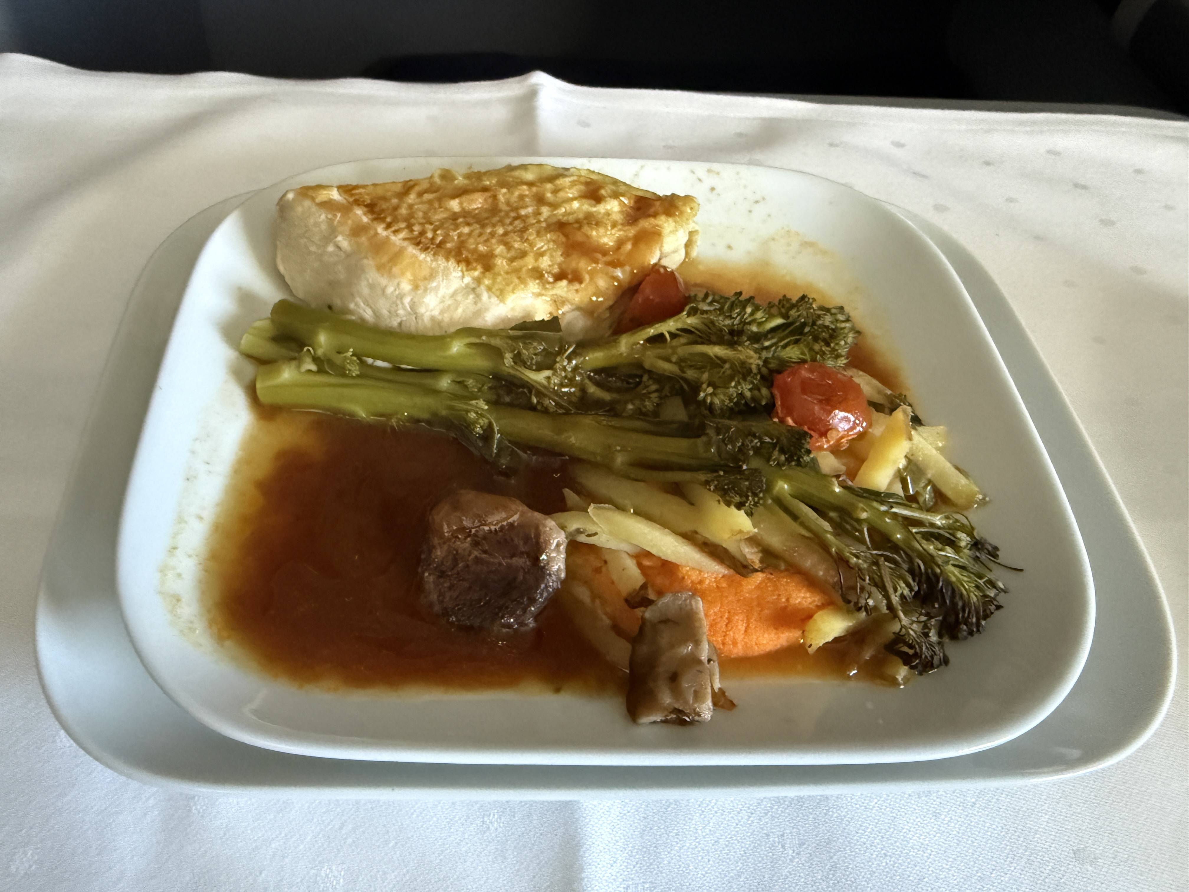 Talíř s kuřecím prsem, brokolini, zeleninou a omáčkou na bílém ubrusu.