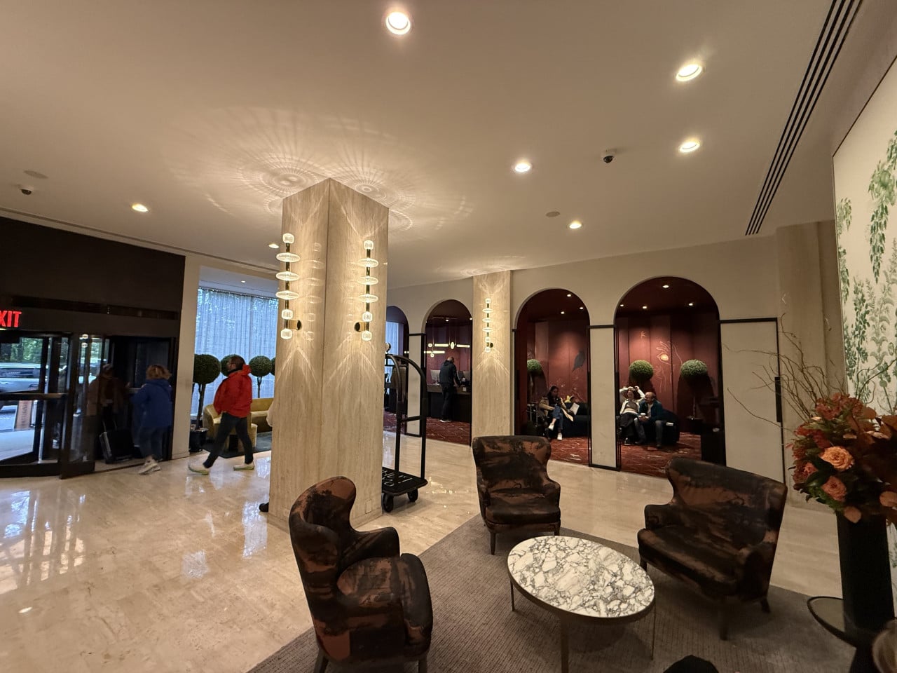 Hotelová lobby s křesly, recepcí, hosty a dekorativními svítidly u vstupu