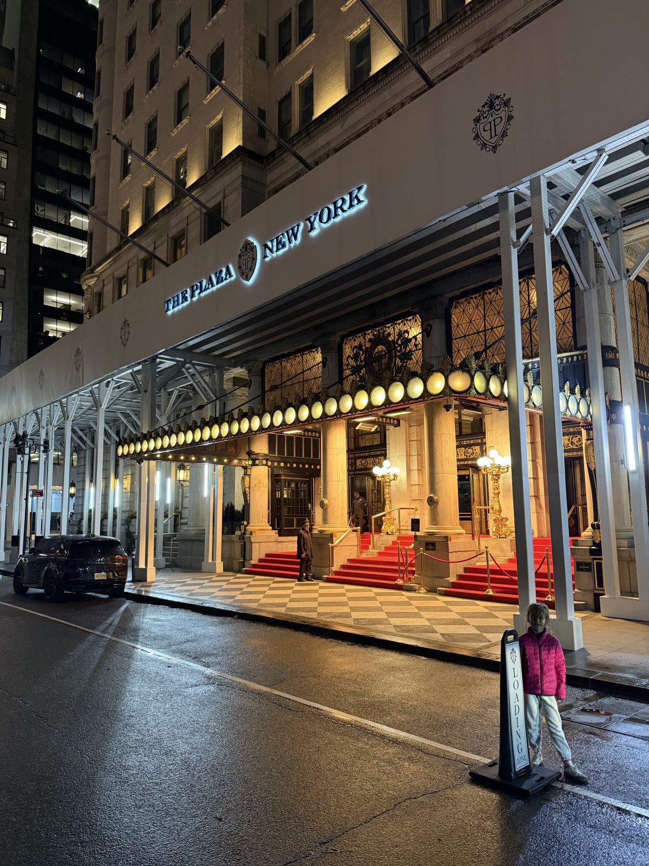 Noční vchod hotelu The Plaza New York s červeným kobercem a dítětem u cedule Loading Zone