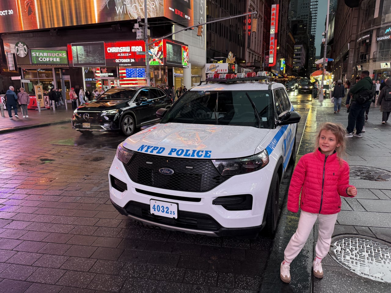Dívka v růžové bundě stojí u policejního auta NYPD na Times Square v dešti.
