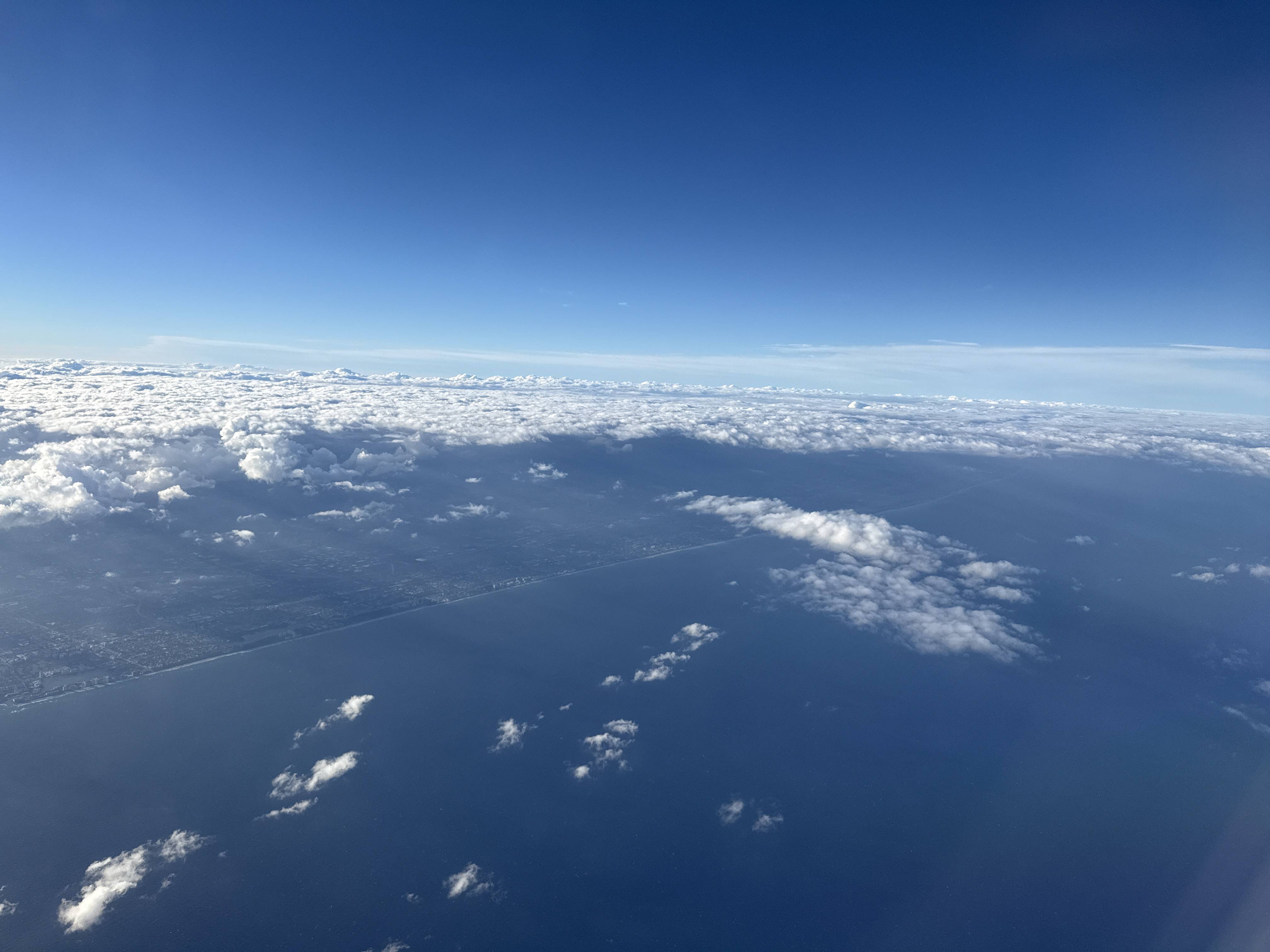 Pohled z letadla na pobřeží, moře a roztrhanou oblačnost pod modrou oblohou