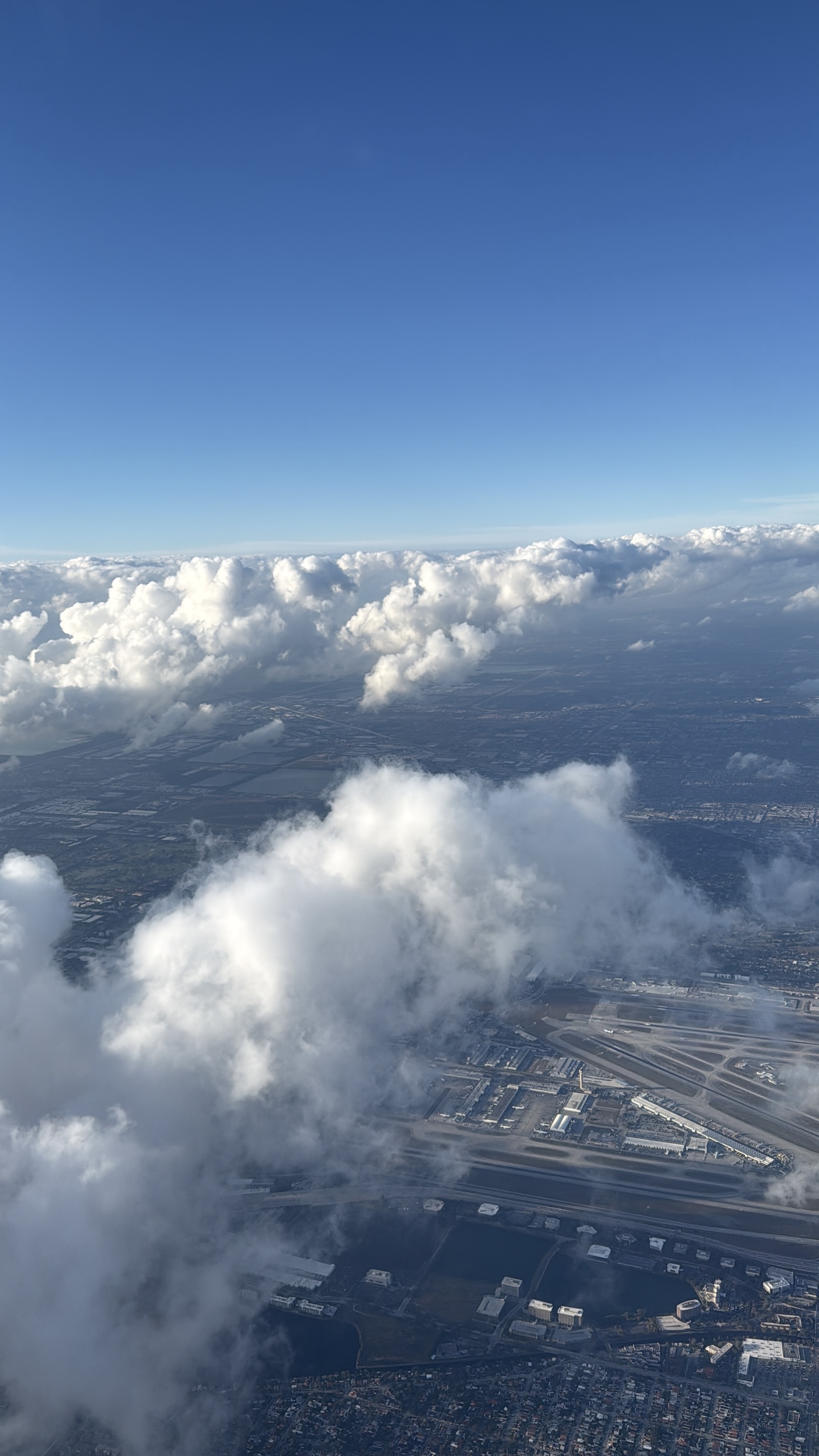 Pohled z letadla na mraky, modrou oblohu a letiště pod sebou